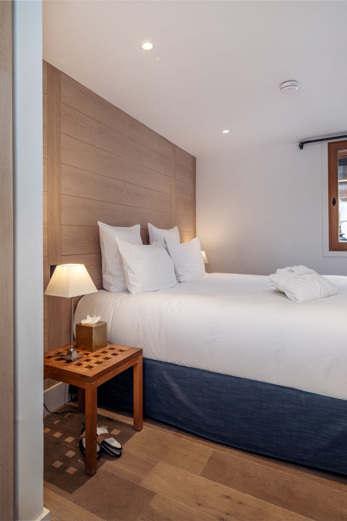 A modern bedroom featuring a neatly made bed with white bedding and a dark navy blue bed skirt, set against a light wood-paneled headboard. A matching wooden nightstand with a lit table lamp sits beside the bed.