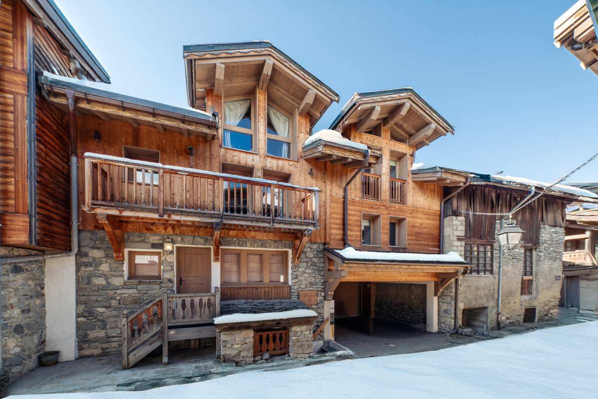 Aerial view of a traditional alpine chalet with wooden balconies, stone foundations, snow on the roof, and a clear blue sky.