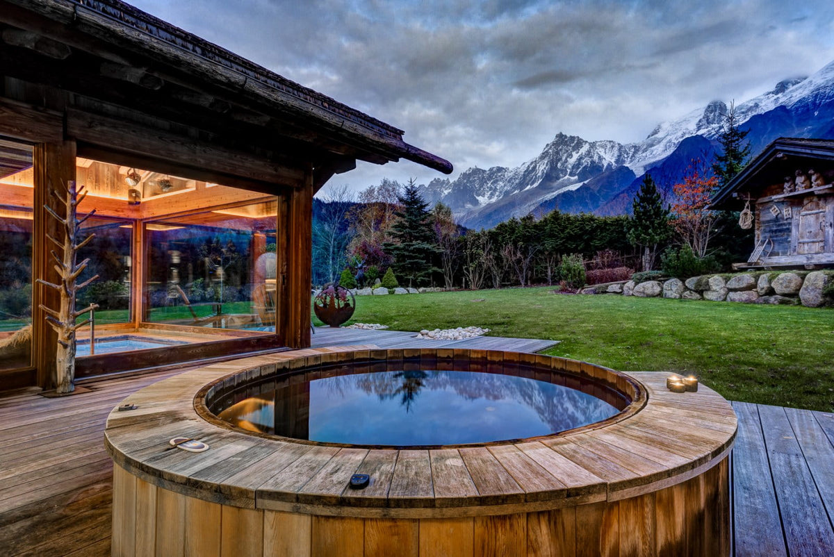 A round wooden hot spring or outdoor spa pool on a deck, set in a luxurious mountain chalet with a green lawn and snow-capped peaks under a cloudy sky.