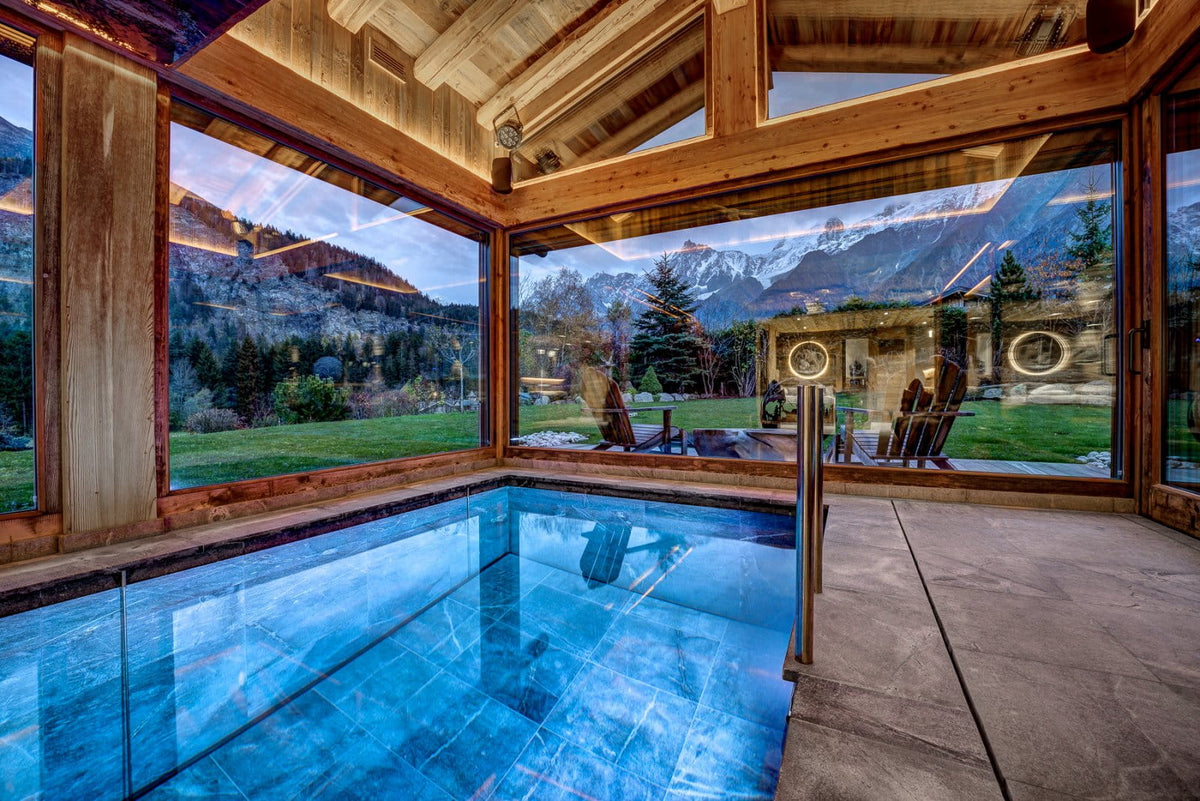 A luxury indoor mountain pool with clear glass walls, showcasing a scenic view of snow-capped peaks and green slopes outside, framed by warm wood paneling and ceiling beams.