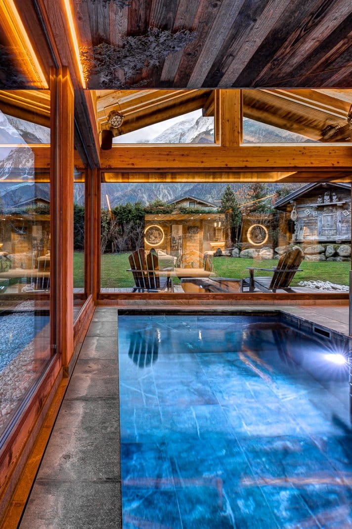Luxury modern chalet interior with a glass-walled indoor pool, warm wood ceiling beams, and an outdoor mountain landscape visible through the windows.