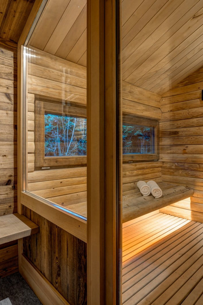 A luxurious wooden sauna interior with a wood-bench, glass sliding door, stone wall, and a tall metal sauna stove on a wood-plank floor.