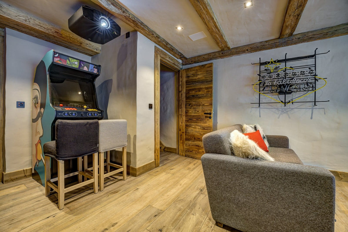 A cozy, rustic living room featuring a gray fabric sofa with a red and white throw pillow, a vintage arcade machine mounted on the wall, and two bar stools. The room has exposed wooden ceiling beams, light-colored plaster walls, and light wood flooring.