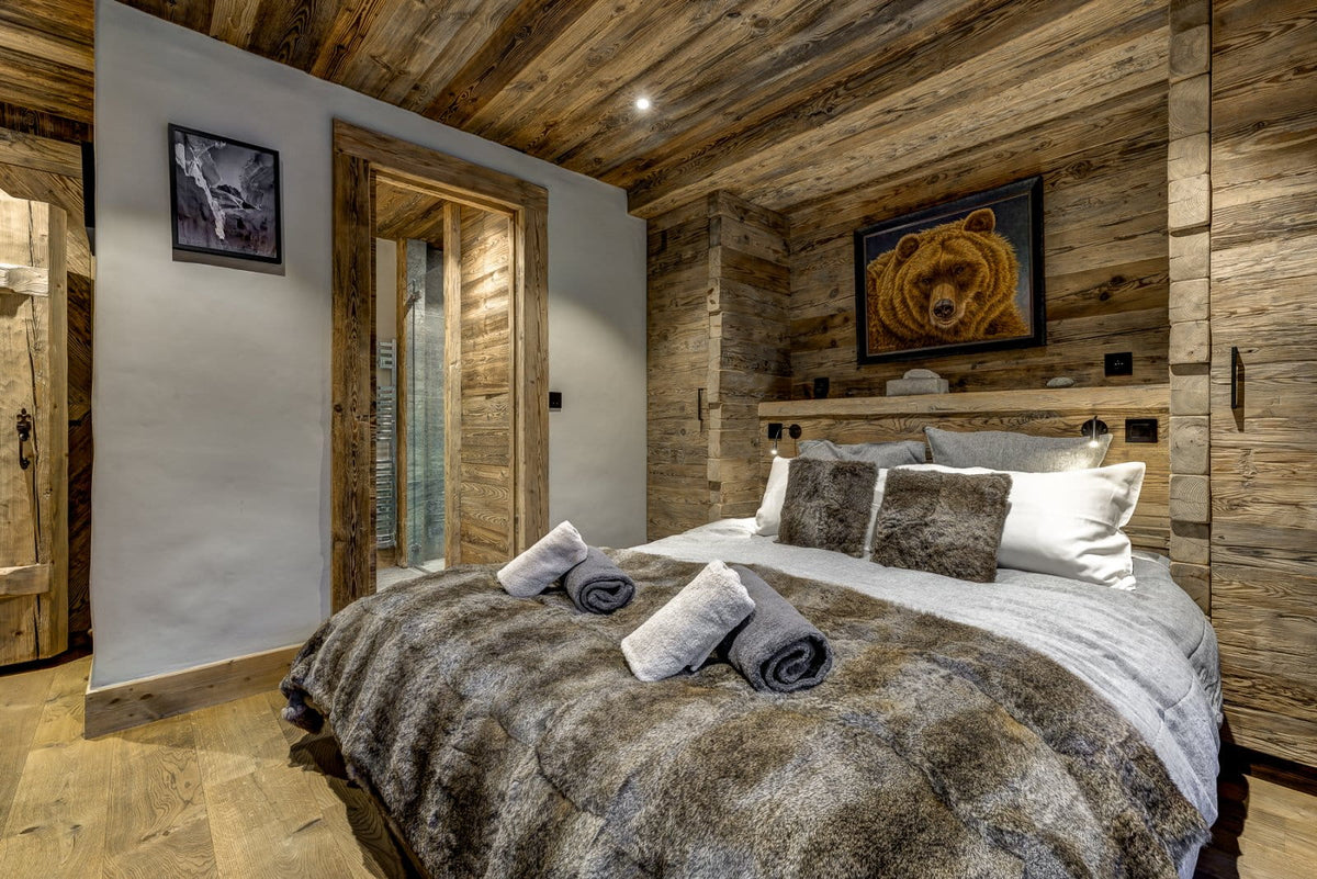 A rustic bedroom with wood-paneled walls and ceiling, featuring a bed dressed in gray and white linens with rolled towels, a framed bear painting above the headboard, and a glass door in the background.