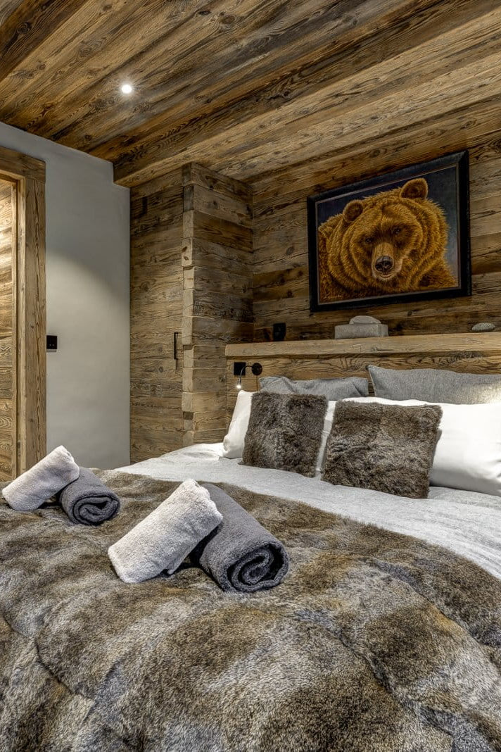 A rustic bedroom with wood-paneled walls and ceiling, featuring a bed dressed in gray and white linens with rolled towels, a framed bear painting above the headboard, and a glass door in the background.