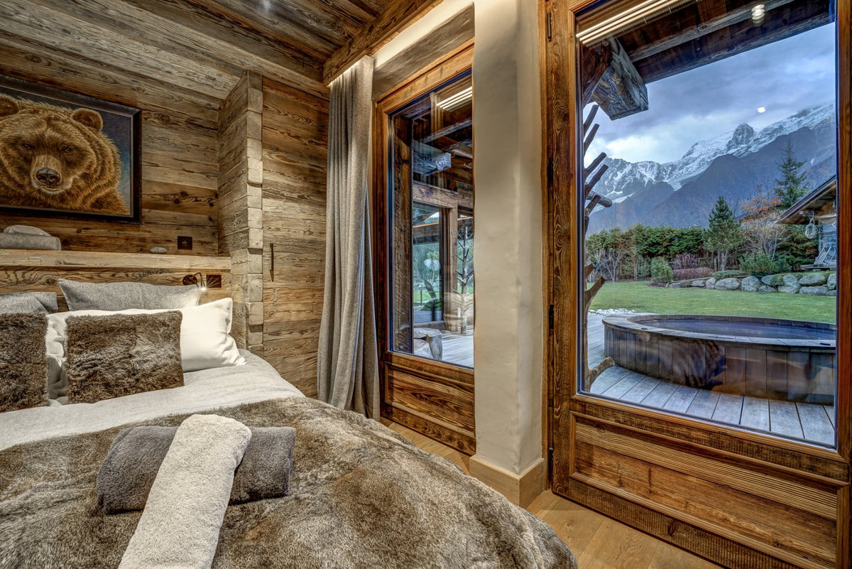 A rustic mountain chalet bedroom featuring a bed with brown and white textured bedding, a wooden headboard, and large windows looking out to a mountain landscape and outdoor hot tub.