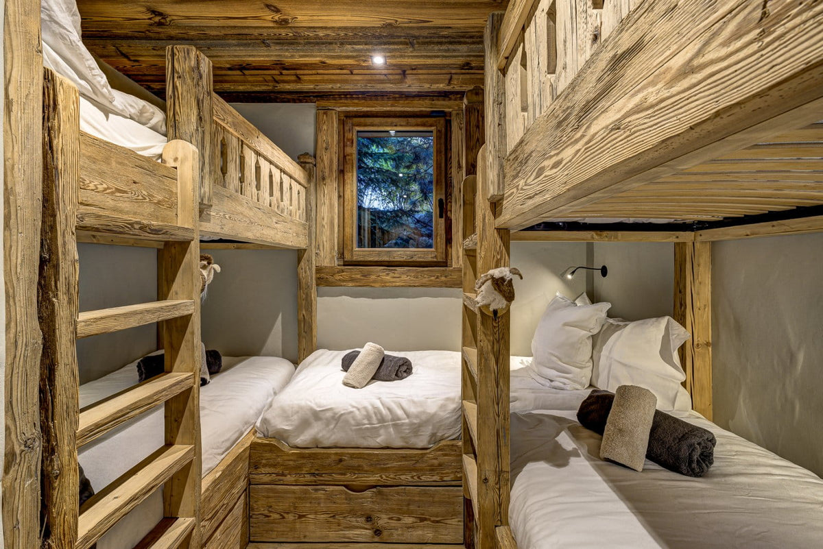 A rustic wooden bunk bed setup in a cabin-style bedroom, with two upper bunks on the left and one lower bunk on the right, all dressed in white bedding. The room has exposed wood paneling and ceiling, a small window looking out onto trees, and bedside wall lights.