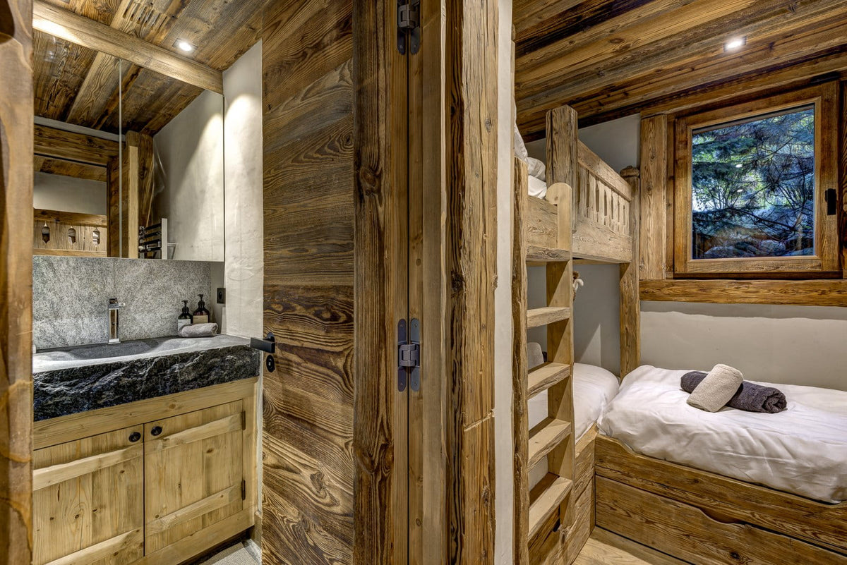 A rustic wooden chalet interior showing a bathroom on the left with a sink and mirror, and a wooden bunk bed on the right with white bedding and a window view of a forest.