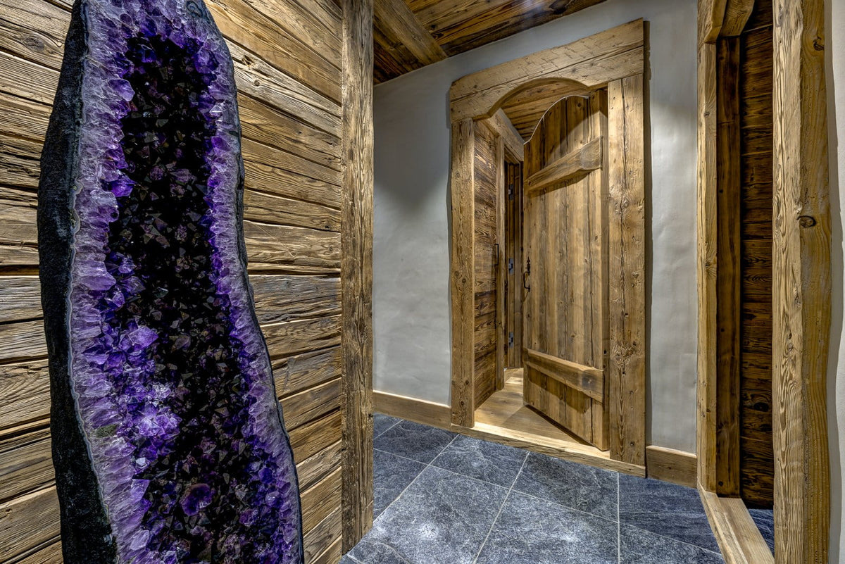 A large purple crystal geode display mounted on a rustic wood-paneled wall in a cabin-style interior with wooden doors and gray stone tile flooring.
