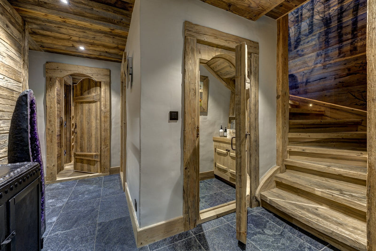 A rustic indoor hallway with exposed wood ceiling beams, wooden doors, dark slate tile flooring, and a wooden staircase on the right. A small vanity area is visible through one doorway, and a dark textured garment hangs on the left edge.