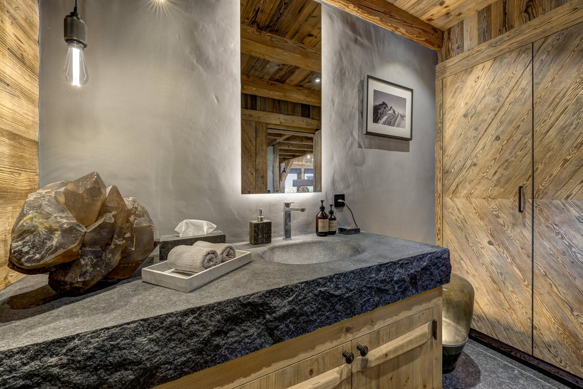 A rustic modern bathroom vanity featuring a dark stone countertop with a built-in sink, a large natural stone sculpture, and a wall-mounted mirror. The cabinetry and storage unit are crafted from warm wood with diagonal grain patterns, and the background wall is a textured light gray plaster.
