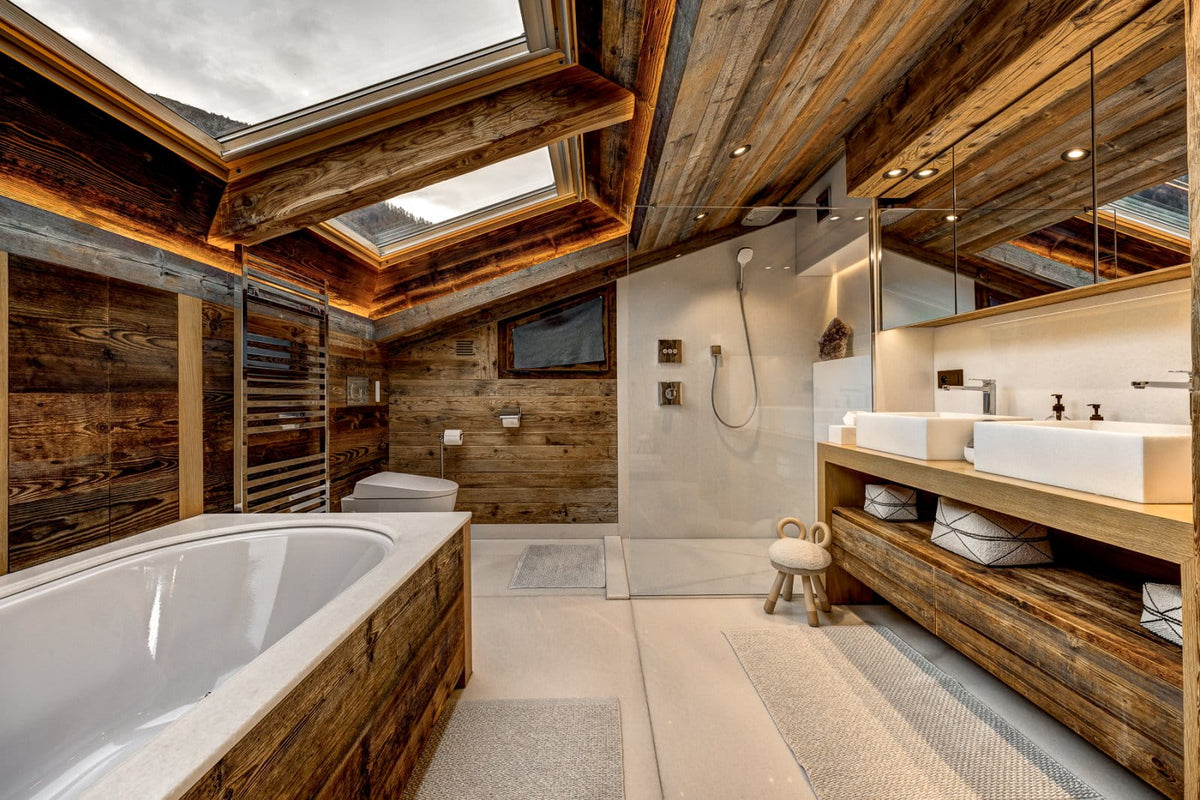 A luxury rustic mountain bathroom featuring a large white freestanding bathtub, a double-sink vanity with white ceramic basins, a glass shower enclosure, and extensive warm wood paneling and ceiling beams under a skylight.
