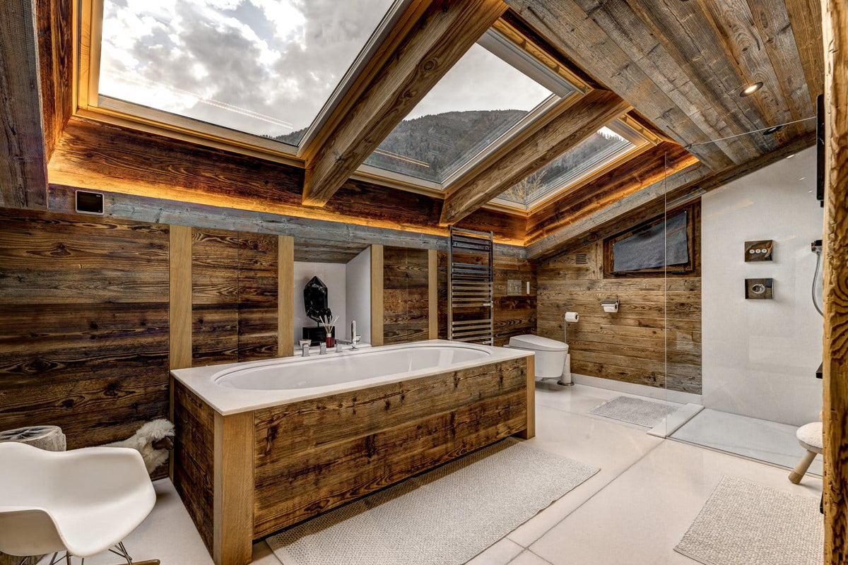 A luxury rustic mountain-style bathroom featuring a large built-in bathtub with a white stone surround, extensive reclaimed wood paneling, a glass shower enclosure, and large skylights looking out to a cloudy sky.