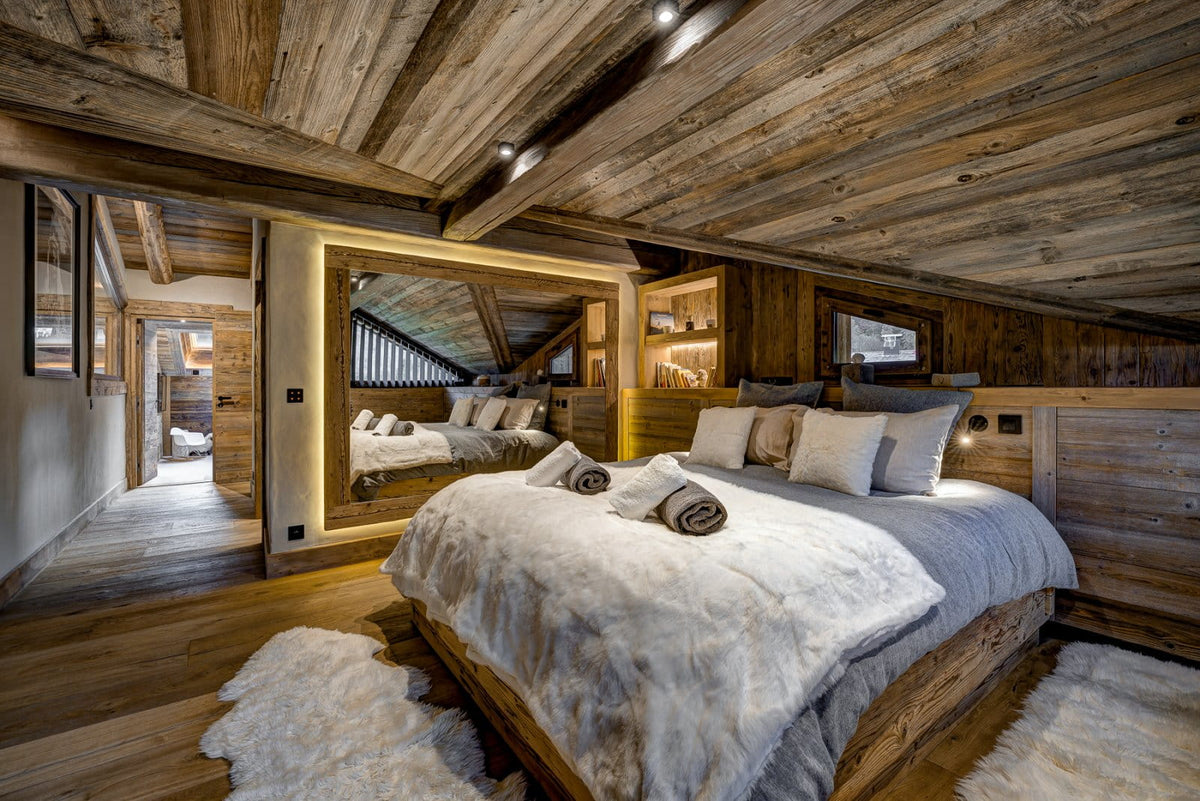 A rustic mountain chalet bedroom featuring a large bed dressed in white and gray bedding, set against a wood-paneled wall with a large mirror reflecting the sloped wooden ceiling. The room is furnished with matching wood flooring and a white sheepskin rug.