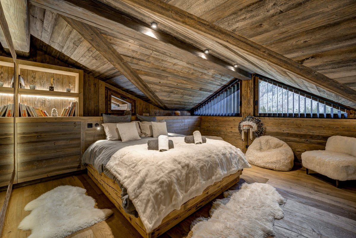 A rustic mountain-style bedroom with a wooden platform bed dressed in cream bedding, set under a sloped wooden ceiling with exposed beams. The room includes a matching wooden chair, a bean bag, and white faux-fur rugs on the floor.