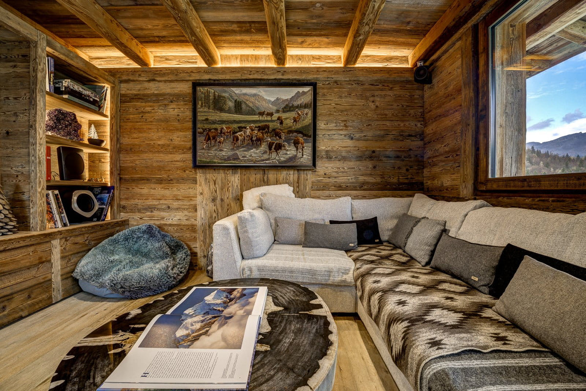 A cozy mountain lodge living room with a large L-shaped sectional sofa upholstered in cream and gray patterned fabrics, set against rustic wood-paneled walls. A framed landscape painting hangs on the wall above the sofa, and a large window on the right shows a scenic mountain view.