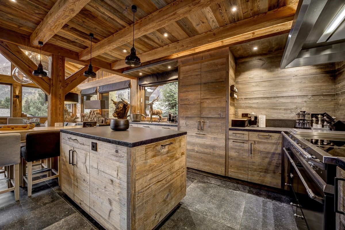 A luxury modern chalet kitchen featuring rustic wood cabinetry, a central island with a dark stone countertop, and a large wood-look backsplash under recessed lighting.