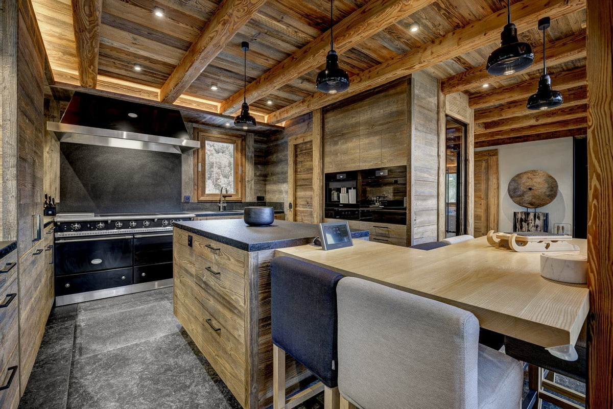 A luxury modern kitchen with rustic wood cabinetry, exposed wooden ceiling beams, black range hood and appliances, a central island with a dark countertop, and a wooden dining table with gray upholstered bar stools.