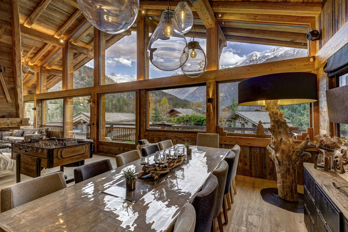 A rustic alpine chalet interior with a long wooden dining table, dark upholstered chairs, large glass pendant lights, and expansive windows overlooking mountains and ski slopes.