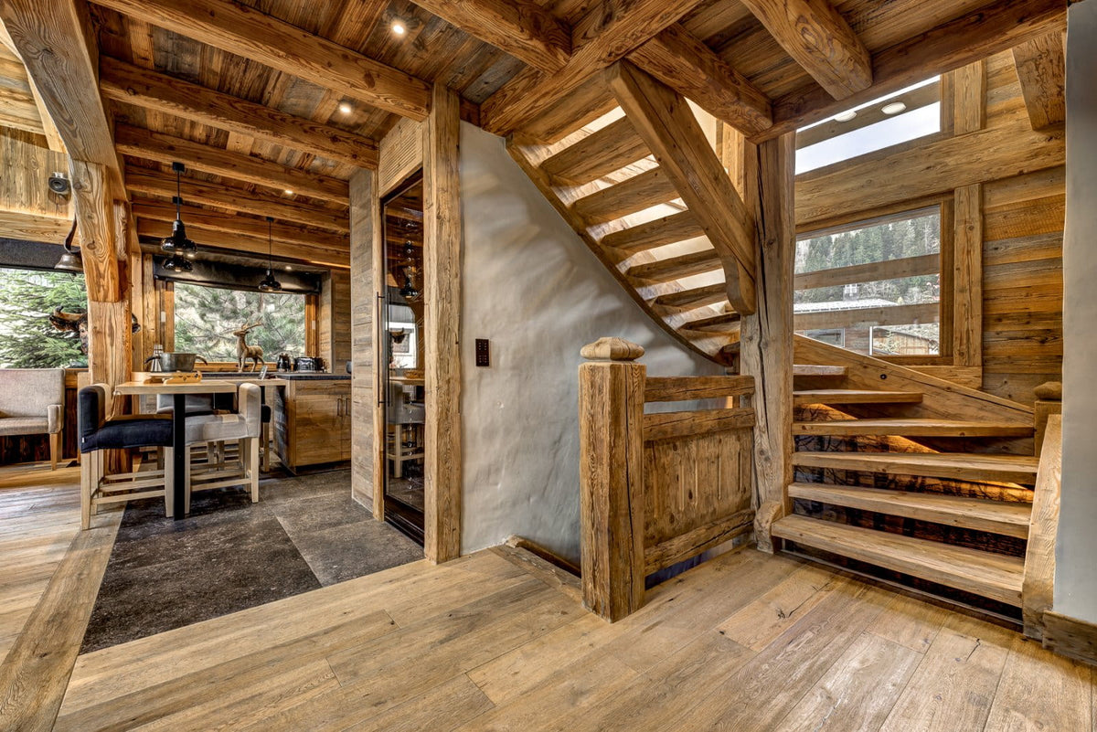 A rustic wooden staircase with a handrail and exposed wood beams in a mountain-style chalet interior, with a dining area and kitchen visible in the background.