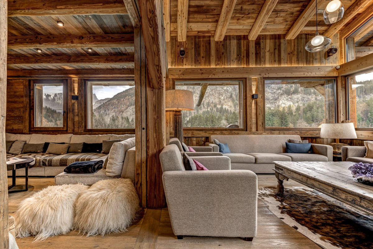 A rustic mountain chalet living room with wood-paneled walls and ceiling, large windows overlooking a forested mountain, beige sofas, a wooden coffee table, and shaggy white poufs.