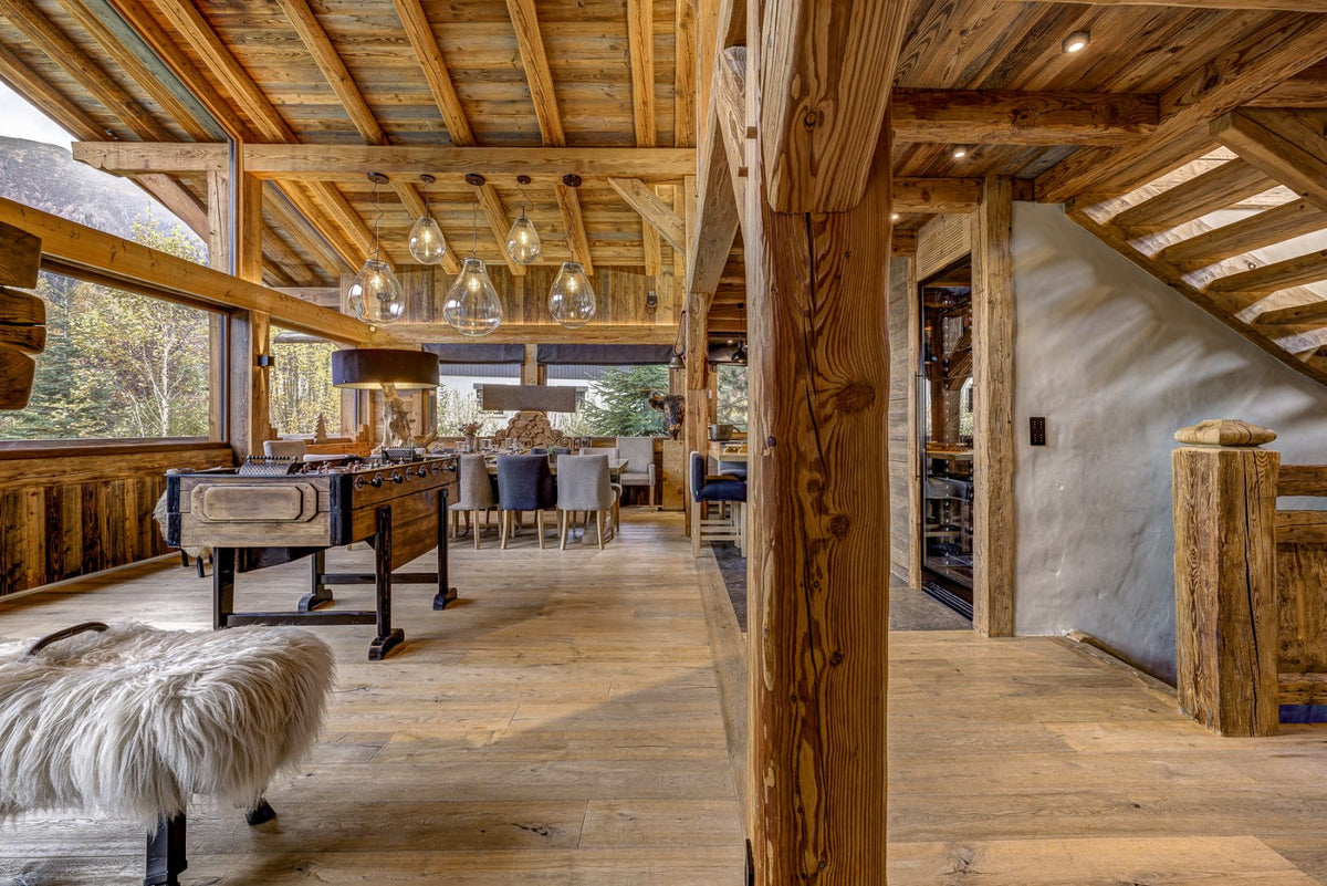 A rustic lodge-style interior with exposed wooden beams, wood floors, a large window view, a dining area with chairs and tables, and a white shaggy bench in the foreground.
