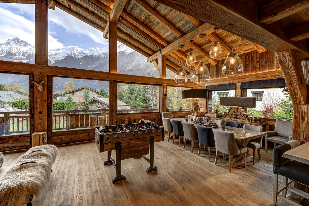 A luxury chalet-style lodge interior with massive wood-framed windows showcasing snow-capped mountains, featuring a wooden foosball table, dining area with gray upholstered chairs, rustic wooden ceiling beams, and warm ambient lighting.