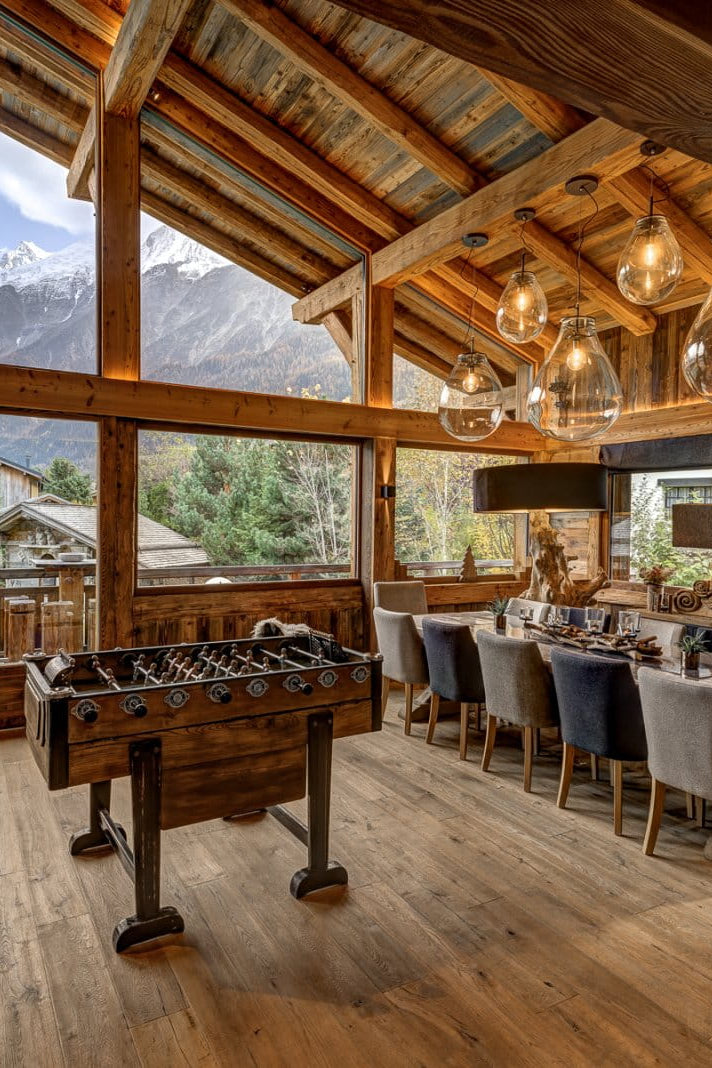 A luxury chalet-style lodge interior with massive wood-framed windows showcasing snow-capped mountains, featuring a wooden foosball table, dining area with gray upholstered chairs, rustic wooden ceiling beams, and warm ambient lighting.