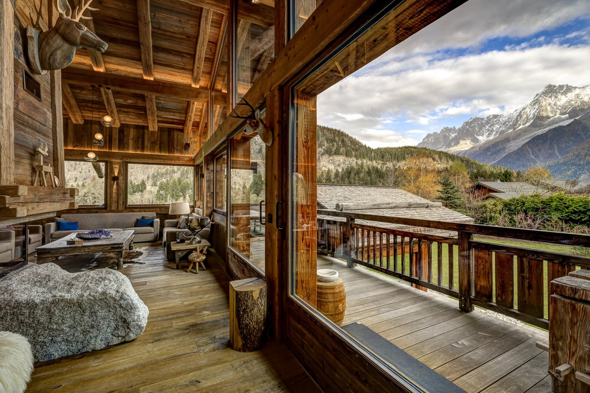 A luxury chalet interior with massive floor-to-ceiling windows overlooking a mountain valley, featuring rustic wood floors, a stone bench, and a decorated wooden ceiling.