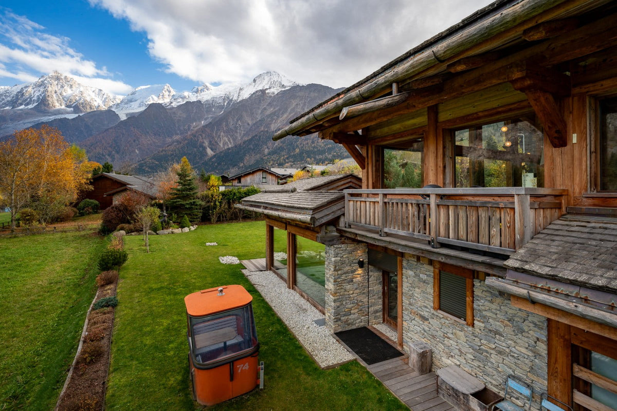 A luxury alpine chalet with stone and wood exterior, featuring a large green lawn, an orange golf cart, and majestic snow-capped mountains under a blue sky in the background.