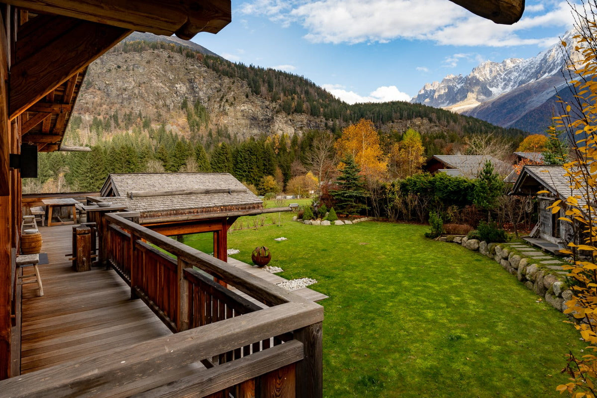 A scenic mountain valley with alpine chalets, a large green lawn, wooden balconies, and snow-capped peaks under a blue sky with clouds.