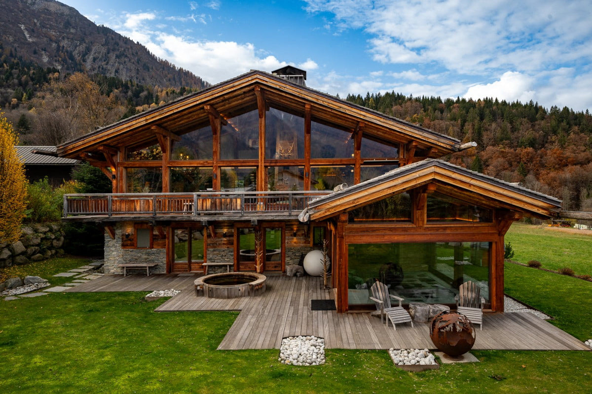 A large modern alpine chalet-style house with extensive wood framing and large glass walls, set on a landscaped lawn with a deck, outdoor seating, mountains, and trees in the background.