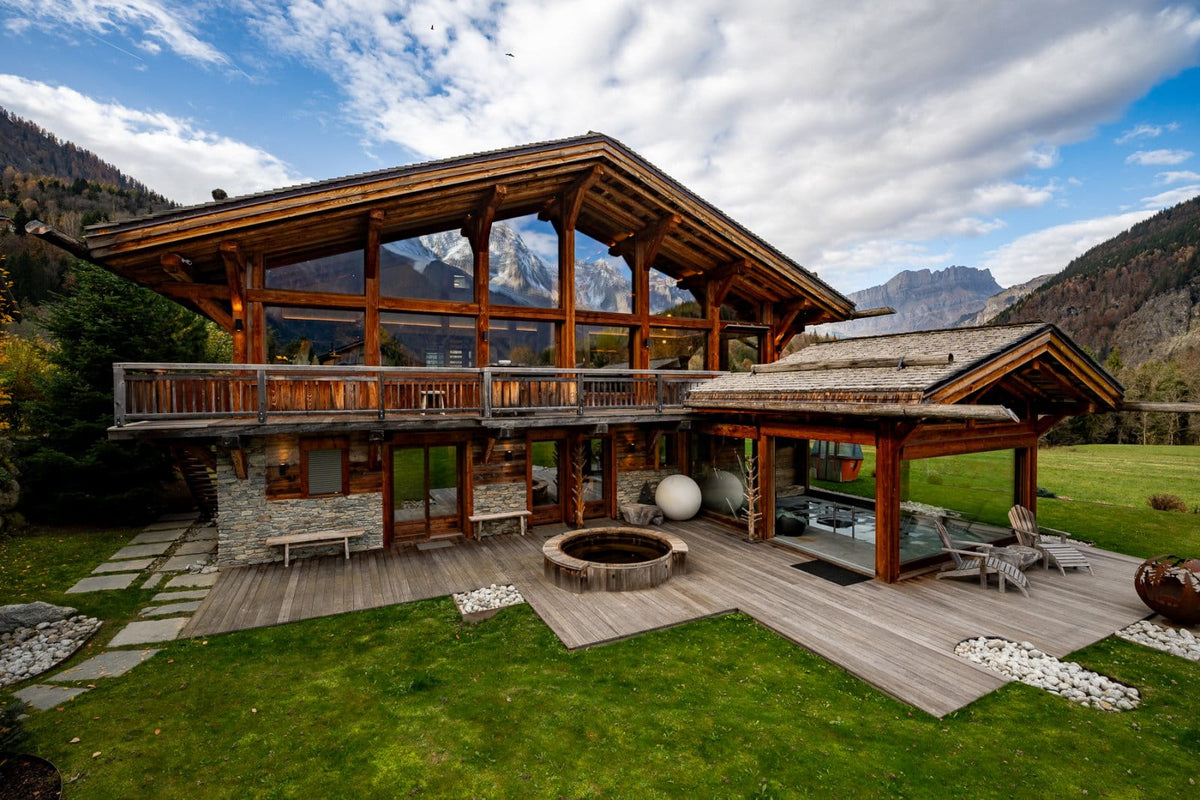 A large modern mountain chalet with a wooden exterior, large glass windows, a wraparound deck, a circular stone fire pit, and a landscaped lawn with mountains in the background.