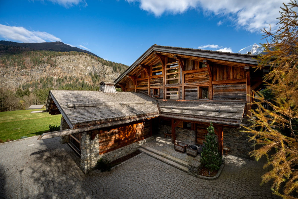 A large rustic alpine chalet with dark wood log walls, a gray slate roof, and stone foundation, set against a backdrop of green fields, mountains, and a bright blue sky.