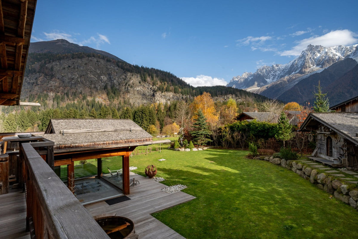 Alpine mountain landscape with snow-capped peaks, forested slopes, and a large green lawn in front of rustic wooden chalets under a blue sky.
