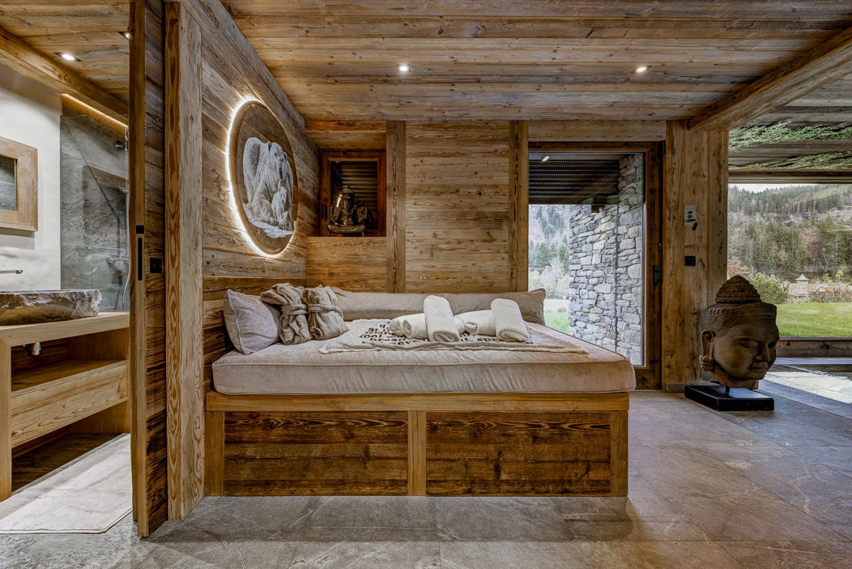 A rustic wooden bed with cream bedding and white towels, set in a modern chalet-style bedroom with wood-paneled walls, stone flooring, and an open doorway looking out to a forested hillside.