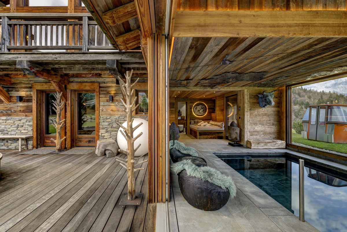 A luxury mountain chalet interior and outdoor deck featuring a wooden deck, stone walls, a modern plunge pool, a large white spherical sculpture, and rustic wooden beams.
