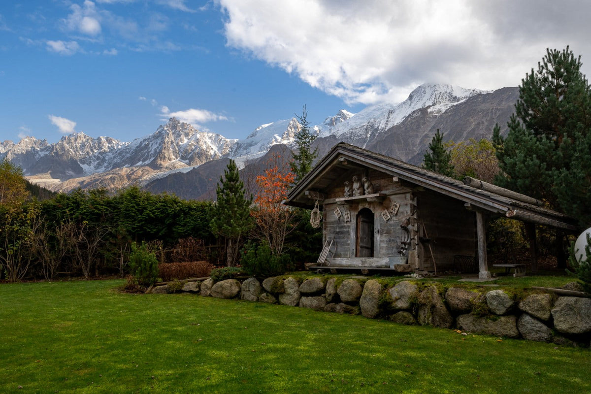 A scenic landscape photograph featuring a rustic carved wooden structure on a stone wall in a green lawn, with a backdrop of majestic snow-capped mountains and a blue sky with white clouds.