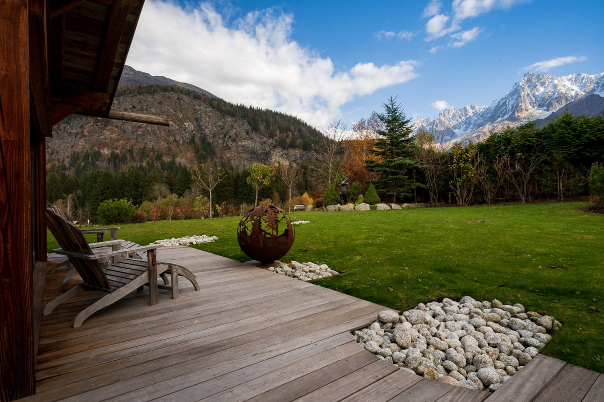A scenic outdoor view from a wooden deck overlooking a large green lawn, with mountains in the background under a blue sky with white clouds. A wooden lounge chair sits on the deck, and the scene is framed by a rustic wooden structure on the left.