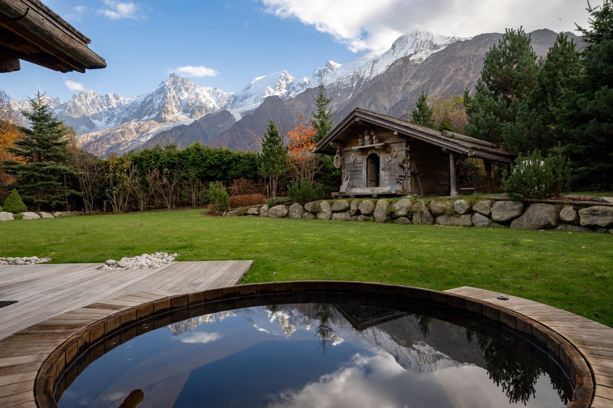 A scenic outdoor setting with a round hot spring pool in the foreground, a stone and wood cabin on a grassy lawn, and snow-capped mountains under a blue sky in the background.
