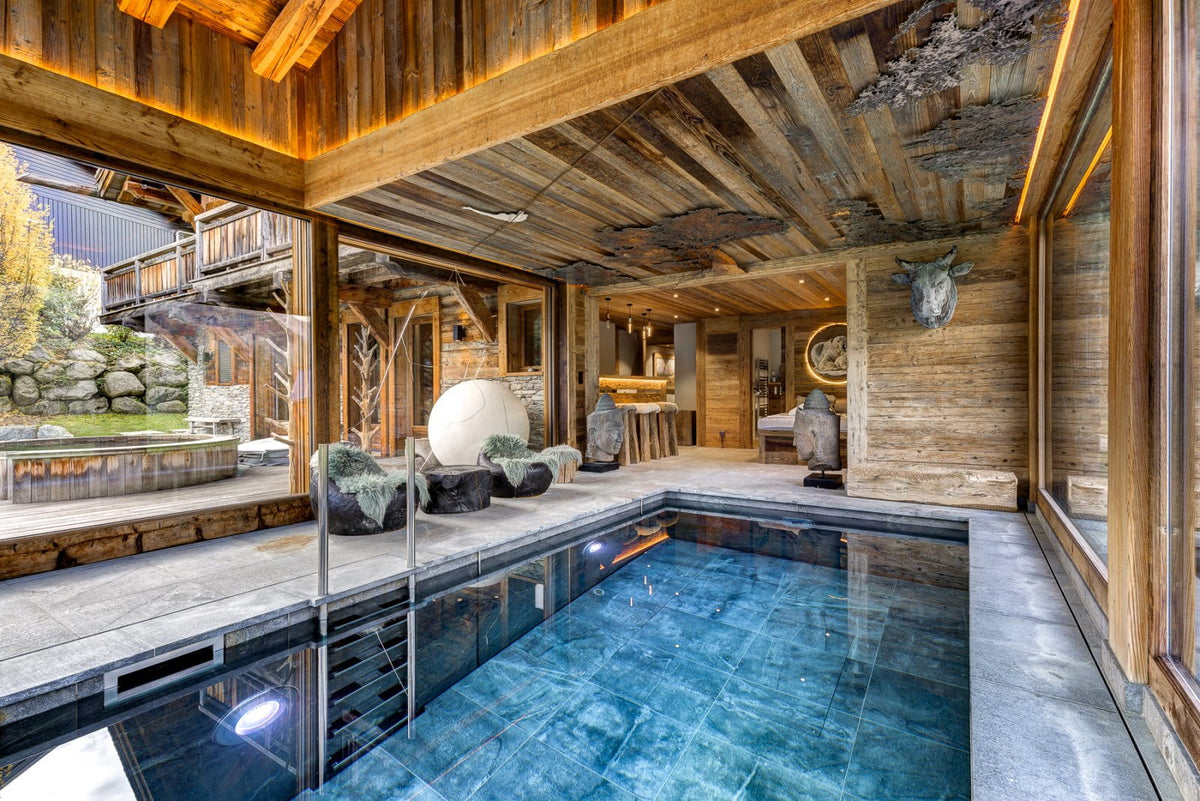 A luxury indoor pool in a rustic wooden chalet, featuring dark blue tiled water, stone flooring, exposed wood ceiling beams, and outdoor views through large glass windows.