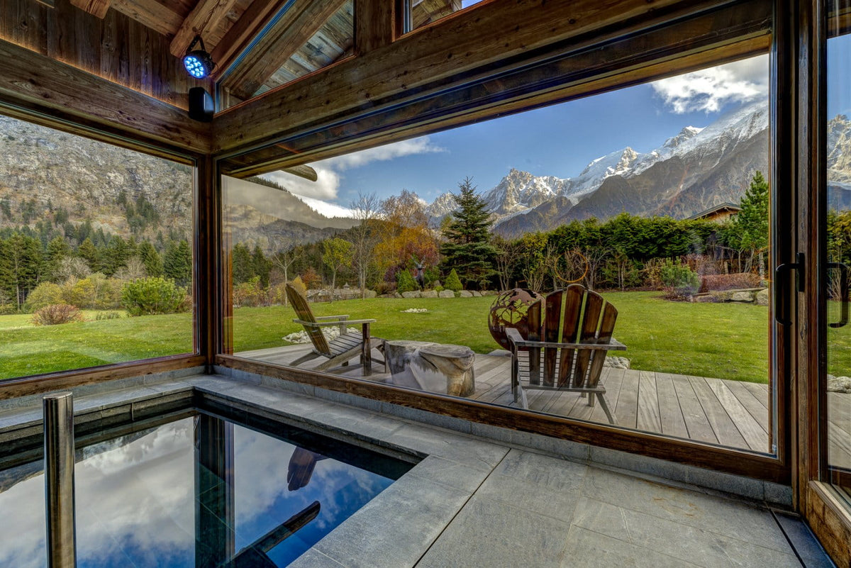 An indoor stone pool overlooking a wooden deck with two Adirondack chairs, set against a scenic mountain landscape with green lawns and snow-capped peaks.