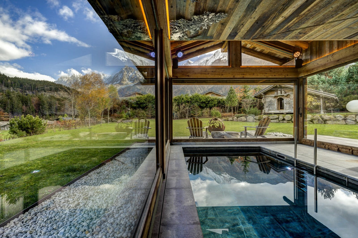 An indoor pool area with large floor-to-ceiling windows overlooking a mountain landscape, grassy lawn, outdoor seating, and rustic buildings. The space features exposed wood ceiling beams, a stone and glass deck, and a rectangular pool with clear blue water.