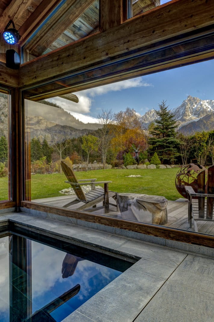 An indoor stone pool overlooking a wooden deck with two Adirondack chairs, set against a scenic mountain landscape with green lawns and snow-capped peaks.