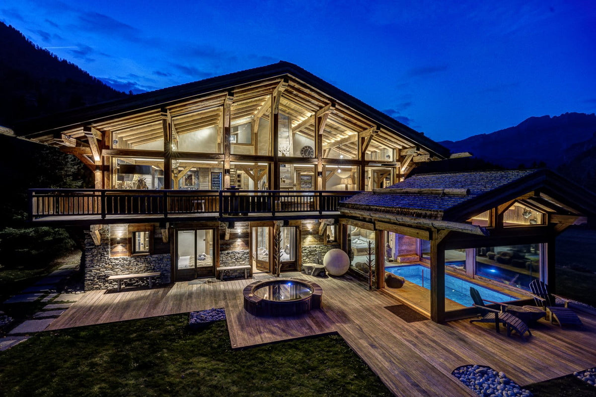 Luxury chalet-style mountain villa at dusk with warm interior lighting, wooden balconies, a stone and wood deck, outdoor hot tub, pool area, and mountains in the background.