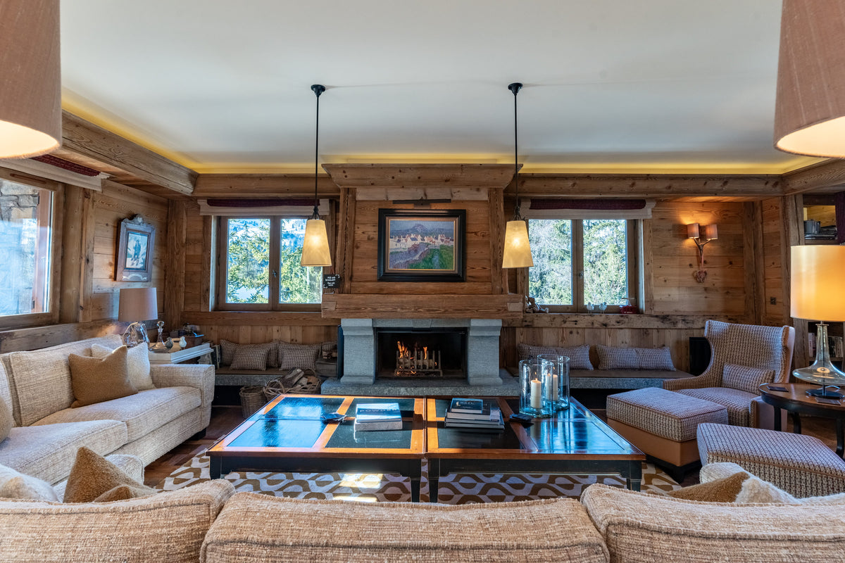 A cozy, rustic mountain-style living room with a fireplace, wood-paneled walls, and multiple beige upholstered sofas arranged around a glass-top coffee table.
