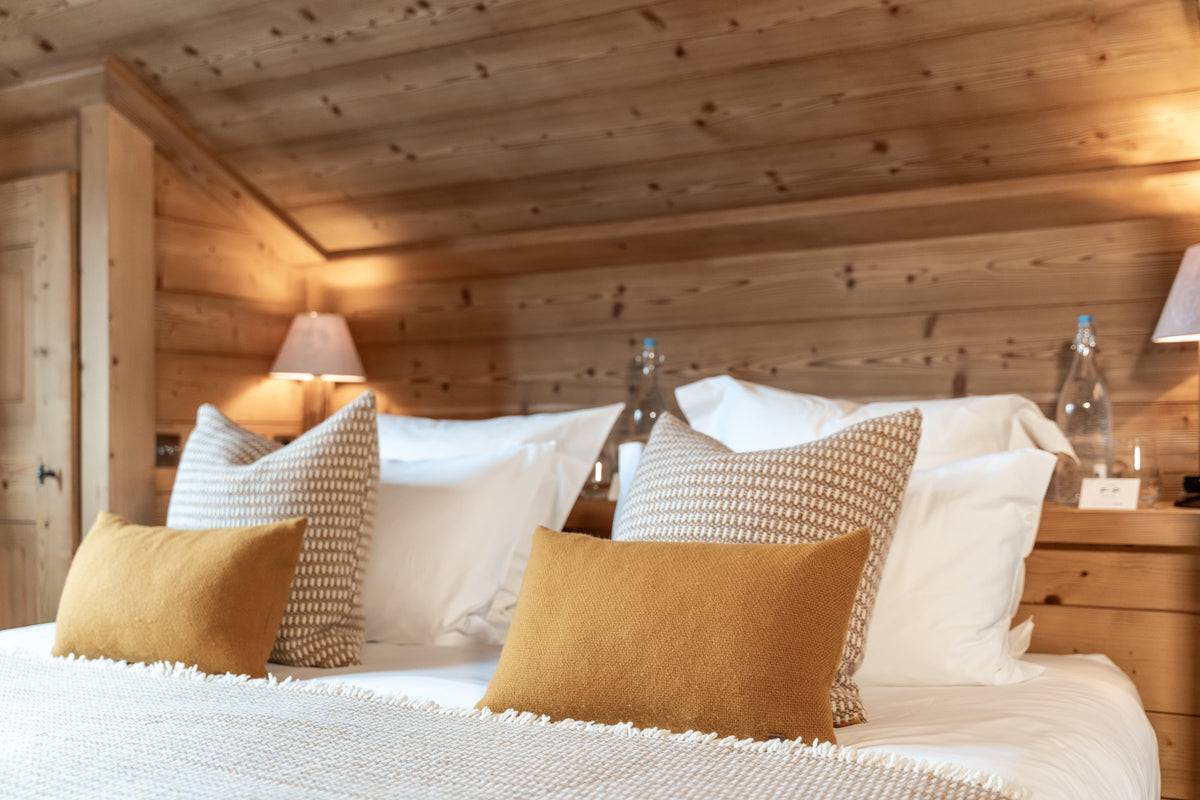 A cozy bedroom scene with a neatly made bed featuring white and patterned pillows, mustard yellow accent pillows, and a textured cream blanket. The room has warm wood-paneled walls and ceiling with bedside lamps and glass bottles.