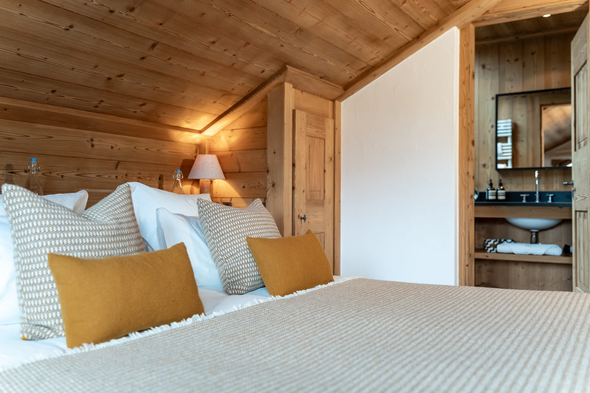 A cozy bedroom interior with a neatly made bed in the foreground, featuring cream textured bedding, white and patterned pillows, and mustard yellow accent pillows. The room has warm wood-paneled walls and ceiling, a wooden door, a bedside lamp, and an open doorway revealing a bathroom with a sink and mirror.