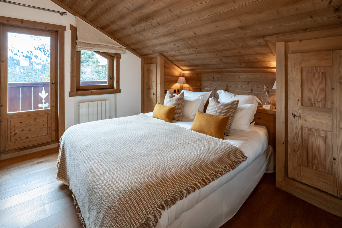 A cozy bedroom with a large bed dressed in white linens and a textured beige throw blanket, accented with mustard yellow pillows. The room features a rustic wooden ceiling, wooden walls, and a matching wooden wardrobe, with natural light coming from a window on the left.