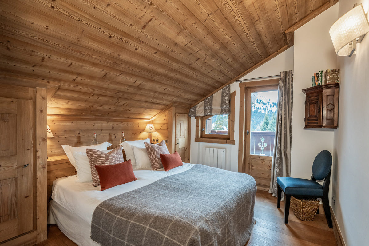 A cozy bedroom with a large bed dressed in white linens, gray plaid bedding, and rust-red accent pillows, set in a rustic wooden chalet with a slanted wood ceiling, window, chair, and wall decor.